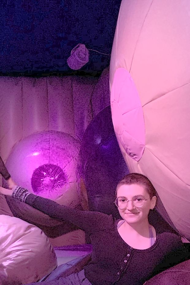 Laine reclining and grinning among oversized soft pink forms under coloured light